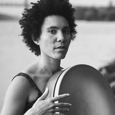 Anaïs Maviel, a thirty-year-old androgynous woman of color, posing with a frame drum in front of the Hudson River in New York City.
